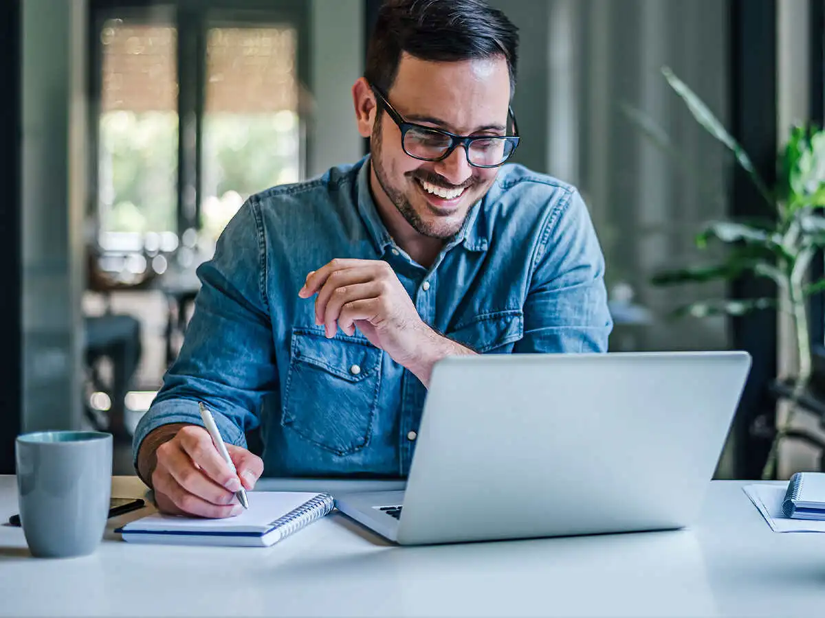 Man writing in a notepad smiling down at a laptop