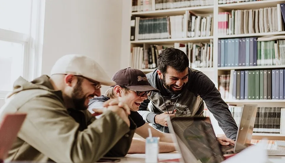 Group of three men in a library smiling down at a laptop