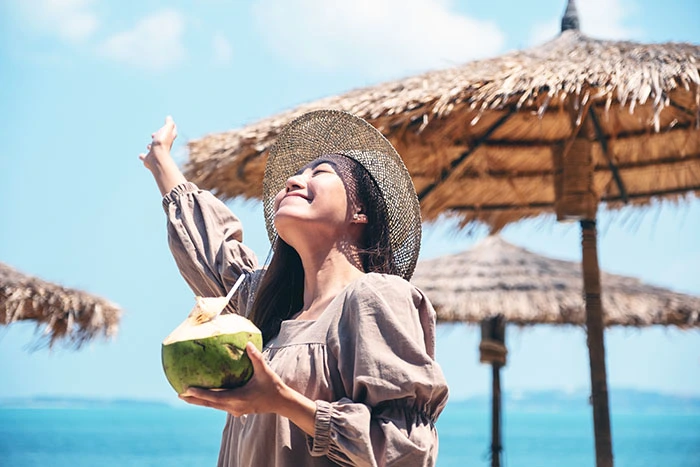 Woman on a beach drinking out of a coconut and smiling up at the sun